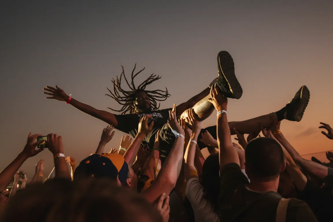 A crowd at a concert lifting a person overhead, hands raised against warm stage light